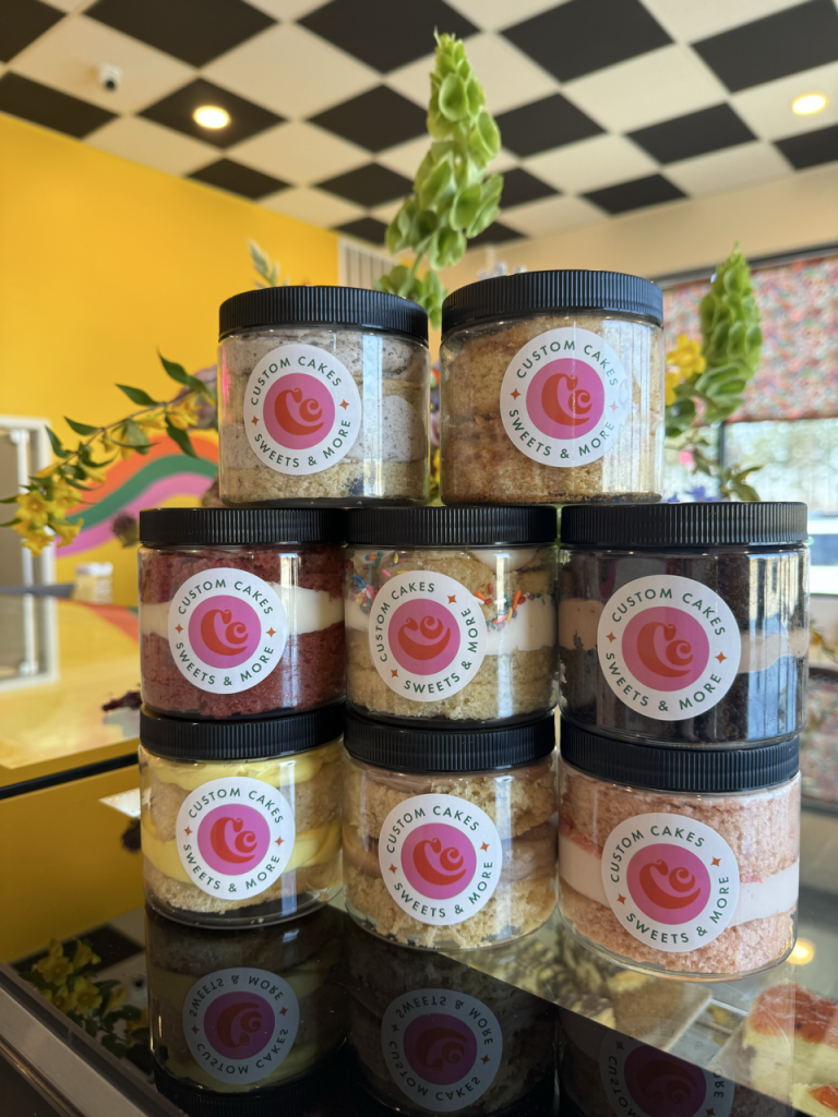 A variety of flavors of cake in a plastic lidded container with the Custom Cake Studio logo on the front, stacked to form a pyramid