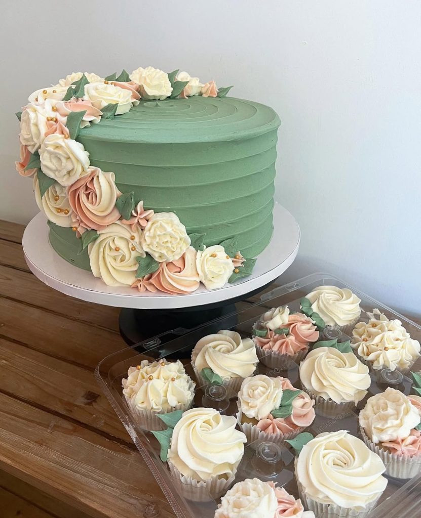 A 1 tier green cake with pink and white florals wrapped around it and cupcakes that match it in a plastic container